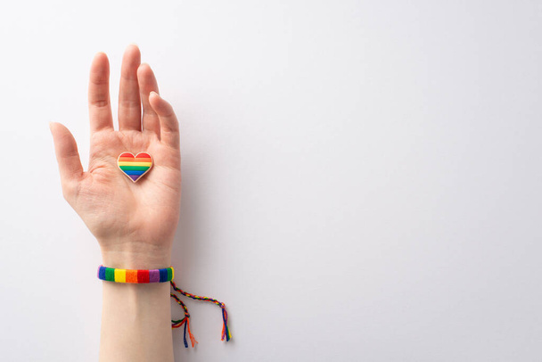 Φωτογραφία πρώτου προσώπου με γυναικείο χέρι σε πολύχρωμο βραχιόλι που κρατά σήμα καρφίτσας σε σχήμα καρδιάς σε λευκό φόντο με χώρο για κείμενο ή διαφήμιση, γιορτάζοντας τον μήνα ιστορίας ΛΟΑΤΚΙ - Φωτογραφία, εικόνα