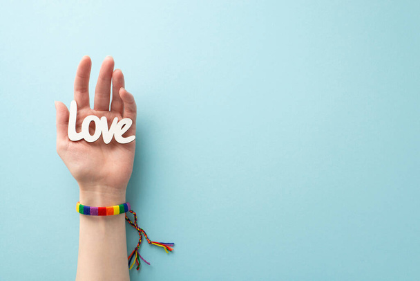 A pastel blue background complements the first person top view image of a woman's hand donning a rainbow bracelet and holding the word "love" on her palm, with space for text or advertising - Foto, imagen