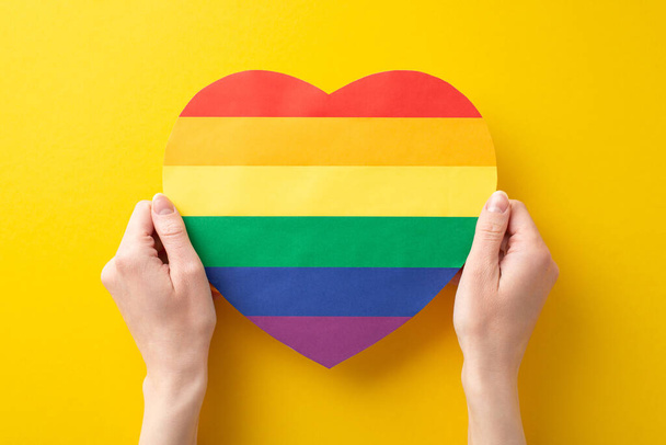 Stehen Sie in Solidarität mit der LGBT-Gemeinschaft mit diesem bedeutsamen ersten peson-Bild von oben, das ein regenbogenherzförmiges Plakat zeigt, das von Frauenhänden auf leuchtend gelbem Hintergrund gehalten wird und Platz für Werbung bietet. - Foto, Bild