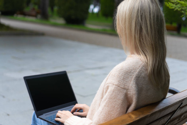 Widok z tyłu młodej blondynki z laptopem na kolanach siedzącej na ławce w parku. Dziewczyna pisze na laptopie w parku - Zdjęcie, obraz