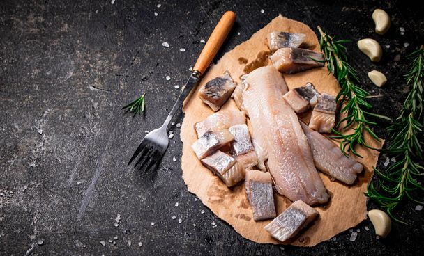 Pieces of salted herring on paper with rosemary and garlic. On a black background. High quality photo - Valokuva, kuva
