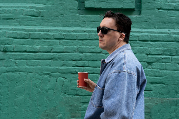 stylish man with dark black hair, wearing sunglasses and blue jeans jacket, standing outdoors next to green wall, holding plastic cup of drink. Street fashion, casual clothes, perfect cover  - Foto, afbeelding