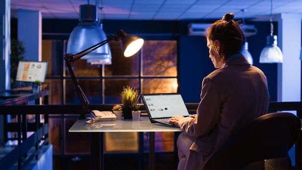 Kaukaska kobieta planuje projekt biznesowy w nocy pracując nad komputerem z analityką internetową. Młody pracownik siedzący przy biurku analizujący statystyki handlu elektronicznego w celu opracowania raportu. Strzał ręczny. - Zdjęcie, obraz