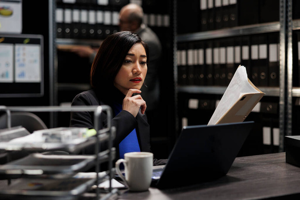 Bureaucratic administration employee checking accountancy budget plan data paperwork on laptop. Asian businesswoman executive in repository filled with document folders and graphical chart reports - Photo, Image