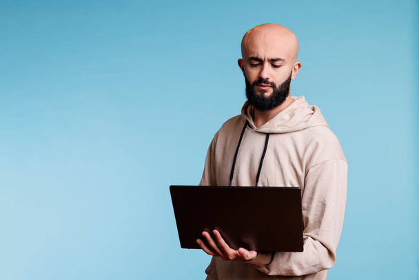 Сосредоточил молодого араба, держащего ноутбук, анализируя код при разработке программного приложения. Запутавшийся человек с озадаченным мышлением выражений во время использования приложения на портативном компьютере - Фото, изображение