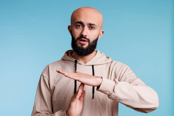 Arab man making time out gesture with hands while looking at camera. Young bald bearded person pausing conversation, showing interruption signal with arms studio portrait - Photo, Image
