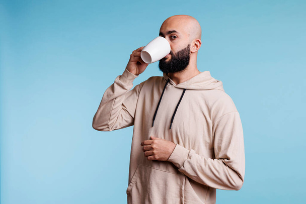 Mladý arabský muž pije kávu z bílého šálku a dívá se do kamery. Arabský plešatý vousatý člověk na sobě ležérní oblečení popíjení čaje, těší horký nápoj z hrnečku studio portrét - Fotografie, Obrázek