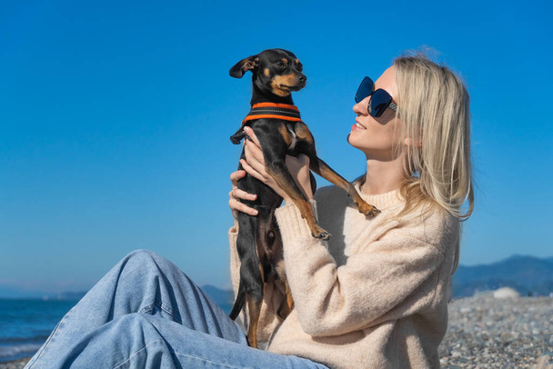 Happy young blonde woman in sunglasses is sitting on the seashore on a sunny day and holding a russian Toy Terrier dog in her arms. Friendship between a dog and a man. - Foto, imagen
