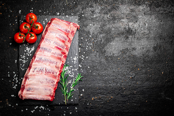 Raw ribs on a stone board with tomatoes and pieces of salt. On a black background. High quality photo - Foto, Imagem