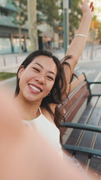 Pretty brunette girl dressed in white top and jeans is smiling making selfie while sitting on bench on cityscape background - Fotoğraf, Görsel