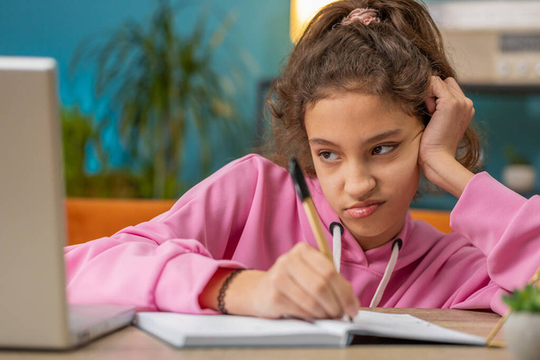 Bored tired Caucasian preteen school girl doing homework exercise online, typing on laptop, makes notes in notebook. Distance learning education. Young child kid at home workplace sitting at the table - Photo, Image