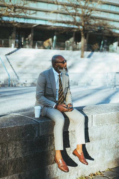 A bald man with a full white-bearded face holding a smartphone, has his cup of coffee next to him, dressed in a cream suit with a yellow striped tie, sitting on limestone steps wearing sunglasses - Photo, image