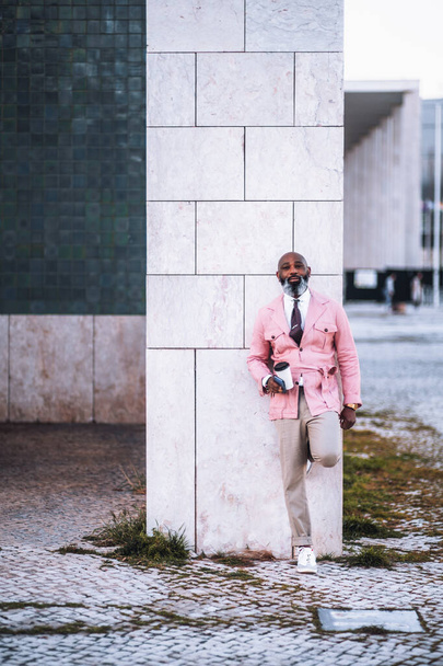 An elegant black bald man, with a long graying beard, dressed in a rose wrap jacket, holds a cup of coffee while leaning against a white marble pillar with an attractive expression on his face - Foto, Imagem