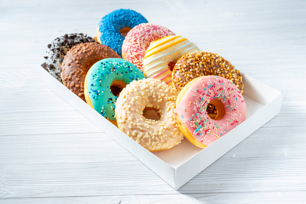 Сладкие пончики разных ярких цветов лежат в коробке на белом столе - Фото, изображение