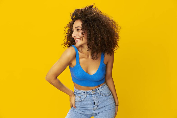 Женщина с вьющимися афро-волосами в синей футболке. Желтые фоновые знаки руками, посмотрите в камеру, улыбнитесь зубами и счастьем, скопируйте пространство. Высокое качество фото - Фото, изображение
