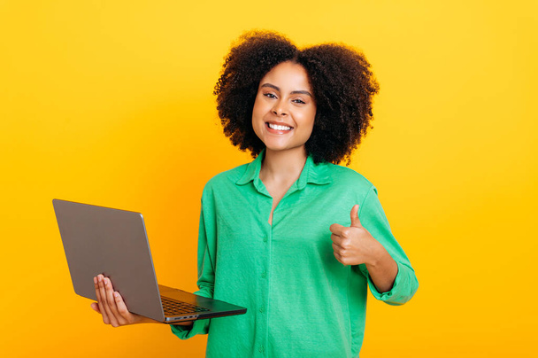 Joyful prachtige Braziliaanse of Afrikaanse Amerikaanse krullende vrouw, in een groen shirt, met een open laptop in de hand, kijkt naar de camera, glimlacht, toont duim omhoog gebaar, staan op geïsoleerde oranje achtergrond - Foto, afbeelding