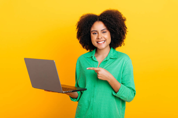 Friendly gorgeous brazilian or african american curly haired woman, in a green shirt, holding an open laptop in hand, points finger at it, looks at camera, smiles, stand on isolated orange background - Foto, Imagem