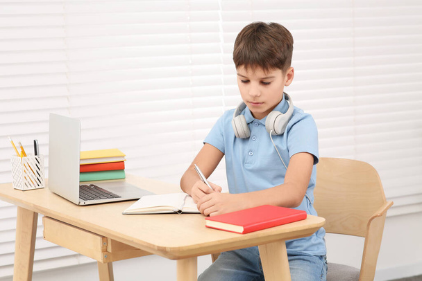 Boy writing in notepad near laptop at desk in room. Home workplace - Fotografie, Obrázek