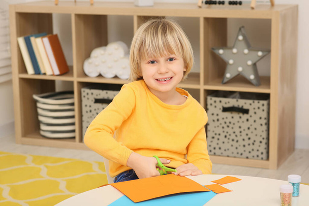 Cute little boy cutting orange paper at desk in room. Home workplace - Φωτογραφία, εικόνα