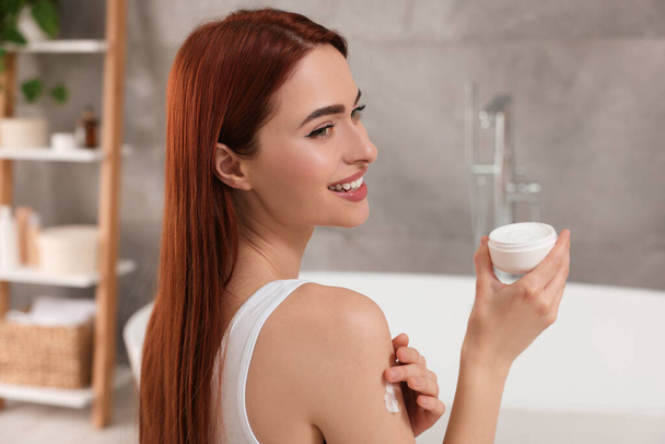 Beautiful young woman applying body cream onto shoulder in bathroom - Valokuva, kuva