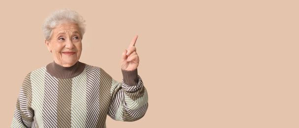 Senior woman pointing at something on beige background with space for text - Фото, изображение