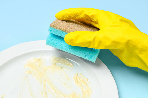 Weibliche Hand in Gummihandschuhen wäscht schmutzigen Teller auf blauem Hintergrund - Foto, Bild