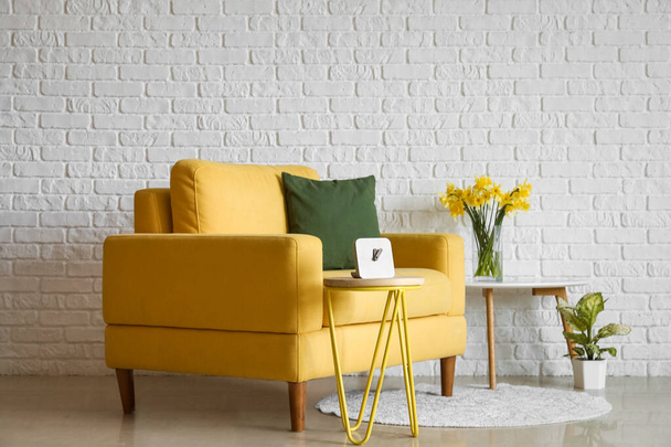 Przytulny żółty fotel i wazon z kwitnącymi narcyzami kwiaty na stoliku w pobliżu białej cegły ściany - Zdjęcie, obraz