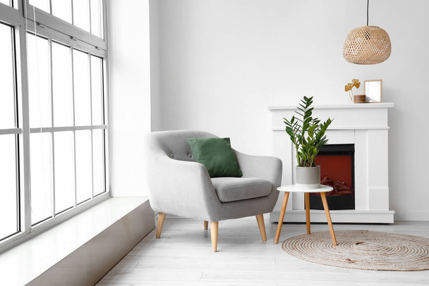 Cozy grey armchair with cushion, fireplace and houseplant on coffee table near big window - Фото, изображение