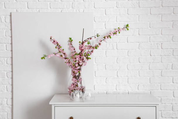 Vase with blooming sakura branches on chest of drawers near white wall - Foto, imagen
