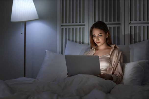 Young woman using laptop in bedroom at night - 写真・画像