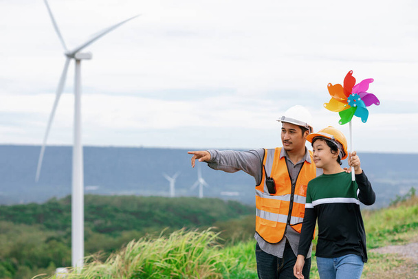 Engineer with his son holding windmill toy on a wind farm atop a hill or mountain. Progressive ideal for the future production of renewable, sustainable energy. Energy generated from wind turbine. - Φωτογραφία, εικόνα