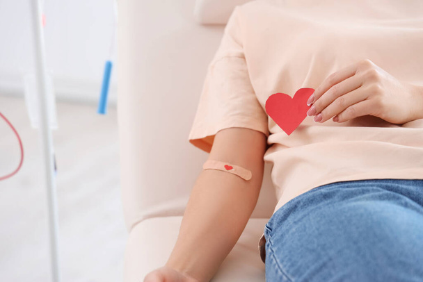Женщина донор крови с прикладным медицинским пластырем и бумажным сердцем в клинике, крупным планом - Фото, изображение