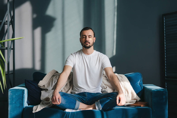 Hombre enfocado meditando en la pose de loto respirando profundo y lentamente usando ropa casual sentado en un cómodo entrenador relajándose en casa. Sentirse libre de estrés. - Foto, Imagen