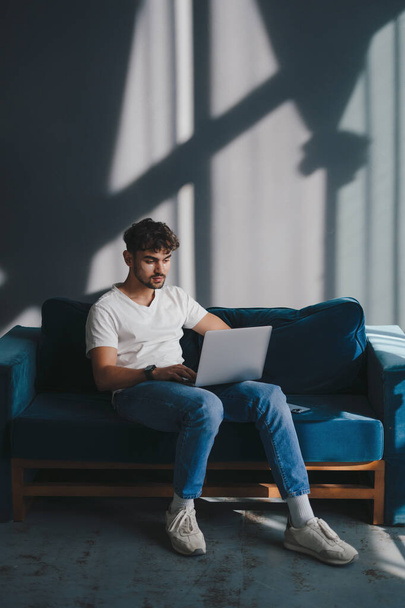 Portret van een jongeman die op de bank zit met een laptop, sms 'jes leest, thuis werkt. Zelfverzekerde man. Internettechnologie. - Foto, afbeelding