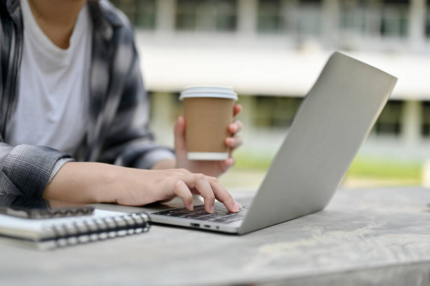 Image en gros plan d'une étudiante asiatique tapant sur un clavier, cherchant de l'information sur Internet, faisant ses devoirs et sirotant un café assis dans un parc du campus. - Photo, image