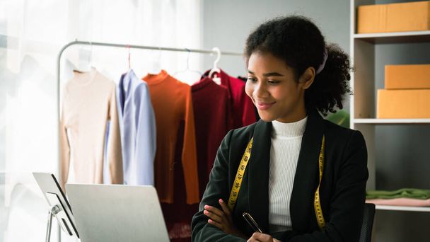 アフリカ系アメリカ人のデザイナーの女性はノートパソコンで顧客の詳細服をチェックし、メモを書く. - 写真・画像