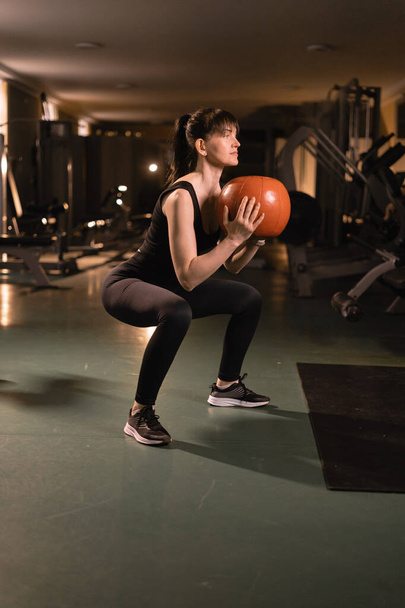 Trainen in de sportschool. Een jonge vrouw hurkt spieren uit door de medicijnbal in de moderne fitnessruimte te houden. Gezondheid en fitness. - Foto, afbeelding