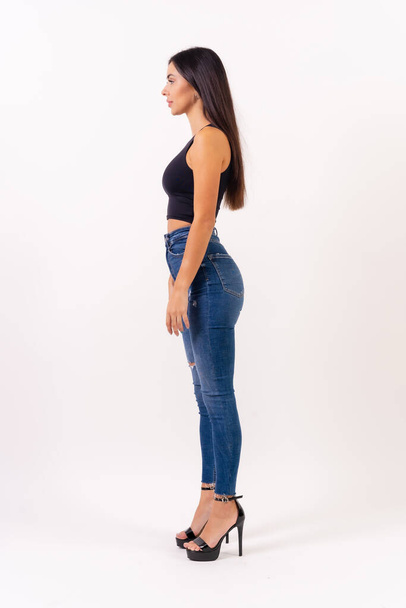Brunette woman in casting photos on a white background, in jeans and a black t-shirt in profile - Fotó, kép