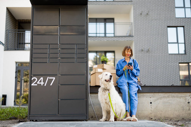 Kobieta siedzi z kartonowymi opakowaniami i białym psem w pobliżu maszyny pocztowej i za pomocą telefonu komórkowego w dzielnicy mieszkalnej. Pojęcie dostawy i stylu życia ze zwierzętami domowymi - Zdjęcie, obraz