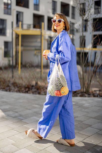 青いパジャマの若い女性がアパートの中庭で新鮮な果物や野菜でいっぱいのメッシュバッグで歩いています。持続可能性、健康的な食品とライフスタイルのコンセプト - 写真・画像