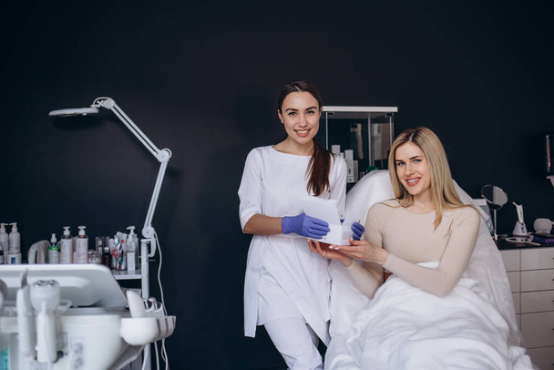 retrato de um cosmetologista com uma seringa na mão e uma cliente mulher sentada em um sofá em uma clínica de cosmetologia. - Foto, Imagem