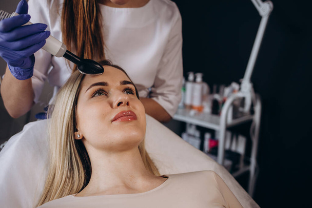 Cosmetologist makes RF lifting for rejuvenation woman face, anti aging cosmetic procedure in beauty spa salon. Beautician makes radio frequency treatment for collagen increasing on female face skin - Zdjęcie, obraz