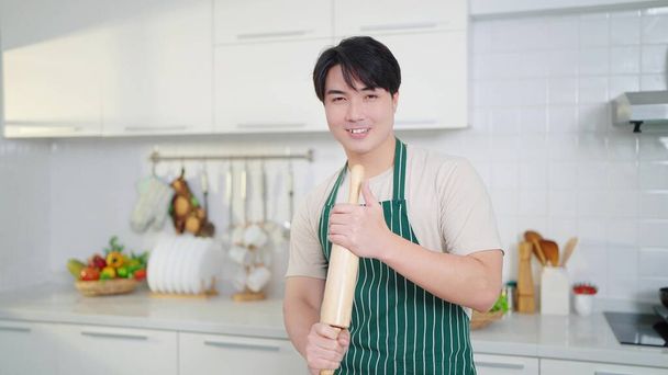 Gelukkig jonge aziatische mannen dragen schort glimlachen voor de camera met een houten rollende pin op handen die staan met vertrouwen in de keuken kamer thuis - Foto, afbeelding