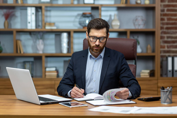 Serious and focused financier accountant on paper work inside office, mature man using calculator and laptop for calculating reports and summarizing accounts, businessman at work. - Photo, Image