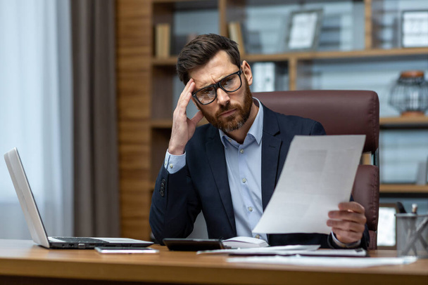 Upset businessman behind paper work inside modern office, mature man with beard reading financial reports and account documents unhappy with results and disappointed with achievements - 写真・画像