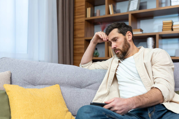 Sad mature man in depression sitting alone at home on sofa in living room. - Photo, image