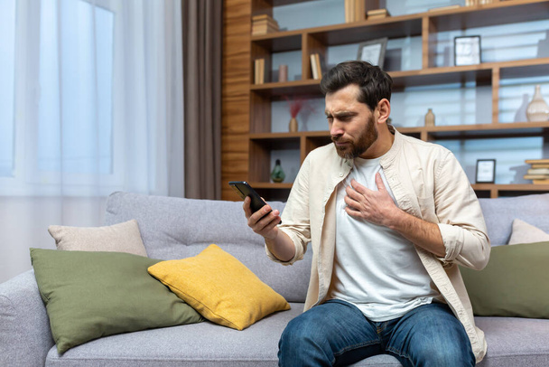 Mature man with beard alone at home reading bad news online on phone while sitting on sofa in living room at home. - Фото, зображення