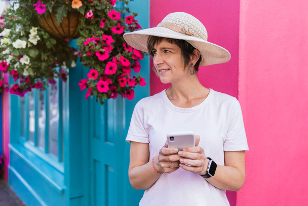 Веселий білявий турист, одягнений недбало, посміхається, набираючи на телефоні повідомлення на задньому плані яскраво забарвлених будинків, прикрашених барвистими рослинами в Ірландії.. - Фото, зображення