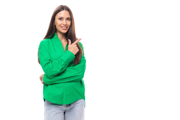 エレガントな緑のシャツでメイクアップした若い白人ブルネットの女性は,空のスペースで壁に向かって彼女の手を指します. - 写真・画像