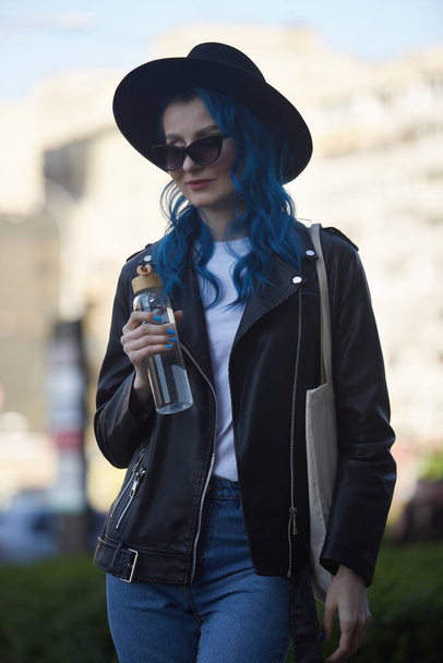 Diverse woman walking outdoor with a glass bottle of water and eco tote bag. Fashionable young adult female with dyed blue hair walks in the city center - Fotoğraf, Görsel